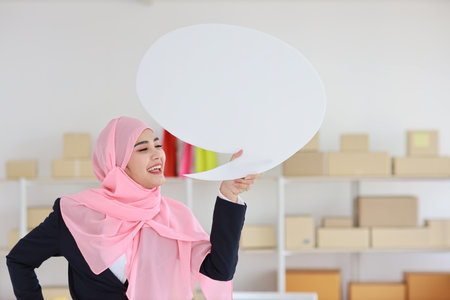 Active asian woman in muslim blue suit standing and holding white speech bubble with online package box delivery background. Startup small business SME freelance girl work at home with happy face.の写真素材