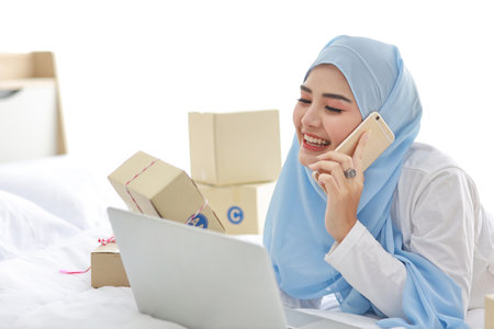 Beautiful and young asian woman in muslim sleepwear with attractive look, lies on bed with computer and online package box delivery. Startup small business SME freelance girl working with mobile phoneの写真素材