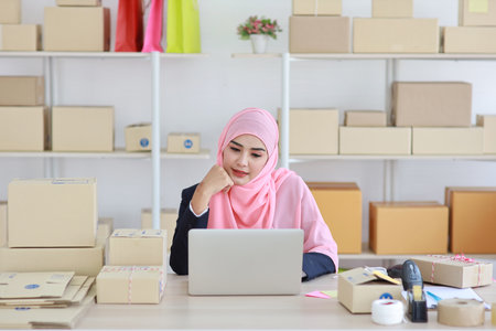 Active asian woman in blue muslim suit sitting and working with computer and online package box delivery. Startup small business freelance working girl look at camera with smilingの写真素材