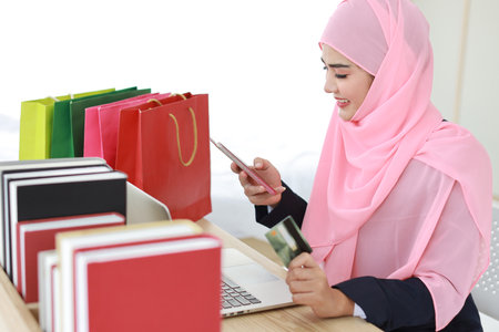 Active smart asian woman in blue muslim suit and pink shaft sitting and holding credit card while using mobile phone for payment or online purchasing. Startup small business girl work at home.の写真素材