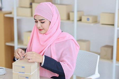 Active smiling asian woman in blue muslim suit sitting and working with online package box delivery. Startup small business SME freelance girl working on computer and mobile phone with happy face.の写真素材