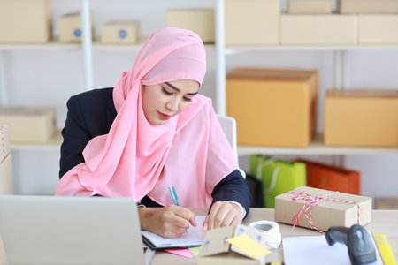 Active asian woman in muslim suit sitting and working with computer, mobile smartphone and online package box delivery. Startup small business freelance working girl checking customer order from bookの写真素材