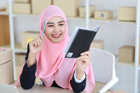 Active asian woman in muslim suit sitting and working with computer, mobile smartphone and online package box delivery. Startup small business freelance working girl checking customer order from bookの写真素材