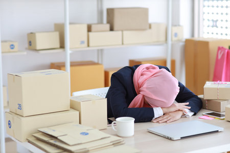 Religious asian woman in blue muslim suit and pink shaft frustrated laid her head down on the table, sit and work on wooden table with computer and package box delivery. Young girl sleep while workingの写真素材