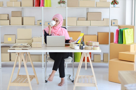 Active smiling asian woman in blue muslim suit sitting and working with online package box delivery. Startup small business SME freelance girl working on computer and mobile phone with happy face.の写真素材