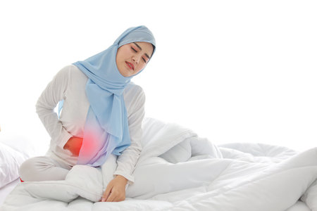 Menstrual pain or stomachache young asian muslim woman sitting on bed. Beautiful girl hold stomach and feeling painful with unhappy face. Healthy conceptの写真素材