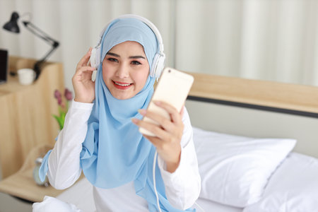 Portrait of beautiful asian female in muslim sleepwear watching online story on mobile phone, lites on bed and connected with wireless internet. Young cute girl with hijab listen music from smartphoneの写真素材