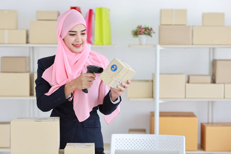 Active asian woman wearing muslim suit standing and using scanner with package box delivery. Startup small business SME freelance girl work at home concept.の写真素材