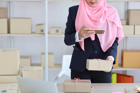 Close up religious asian woman in muslim blue suit standing and taking picture of package box deliver from mobile phone. Startup small business SME freelance girl work at home with happy smiling faceの写真素材