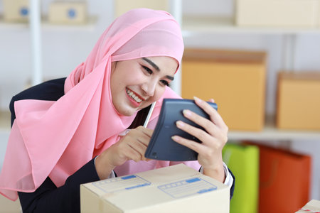 Active smiling asian woman in muslim suit sitting and working with online package box delivery. Startup small business SME freelance girl working on computer and mobile phone with happy face.の写真素材