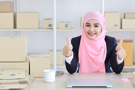 Active asian woman in blue muslim suit sitting and working with computer and online package box delivery. Startup small business freelance working girl showing thumbup with exciting emotion.の写真素材