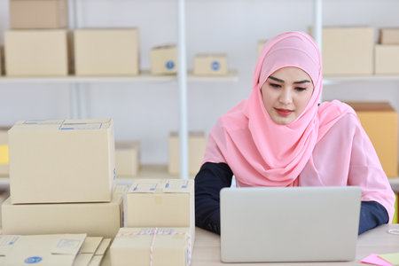 Active asian woman in blue muslim suit sitting and working with computer and online package box delivery. Startup small business freelance working girl look at camera with smilingの写真素材