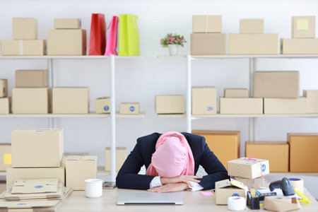 Religious asian woman in muslim blue suit and pink shaft frustrated laid her head down on the table, sit and work on wooden table with computer and package box delivery. Young girl sleep while workingの写真素材