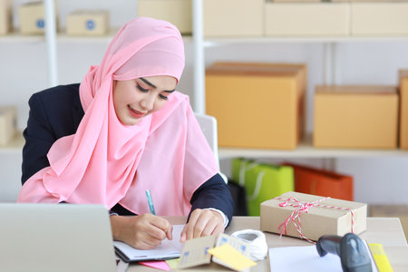 Active asian woman in muslim suit sitting and working with computer, mobile smartphone and online package box delivery. Startup small business freelance working girl checking customer order from bookの写真素材