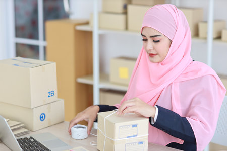 Active smiling asian woman in blue muslim suit sitting and working with online package box delivery. Startup small business SME freelance girl working on computer and mobile phone with happy face.の写真素材