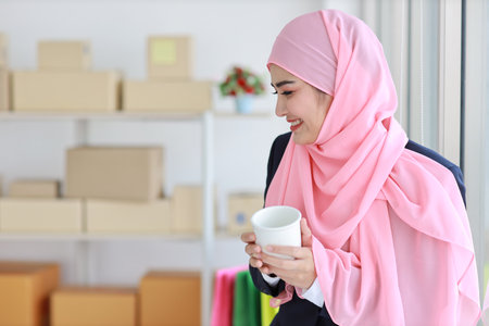 Religious asian woman in muslim blue suit drinking coffee, smiling and pink scarf on head with package box delivery background. Startup small business SME freelance girl work at home with happy faceの写真素材