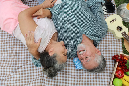 Top view happy asian senior man and woman lying on blanket and having fun on picnic together in garden outdoor. Lover couple eating food and embracing at the park. Happiness marriage lifestyle conceptの素材 [FY310207740962]