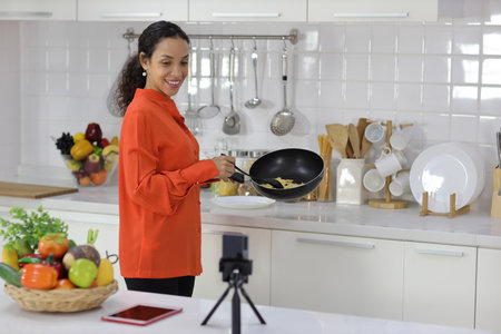 Happy latin woman holding a pan for cooking while checking recipe or learning from internet with mobile phone in kitchen. Young female preparing delicious meal at home. Healthy and lifestyle conceptの写真素材