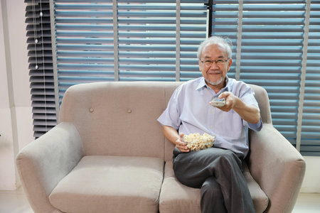 Happy smiling asian senior man sitting on sofa and eating popcorn while having fun with movie rest indoor at home living room. Elderly husband is happiness while watch TVの写真素材