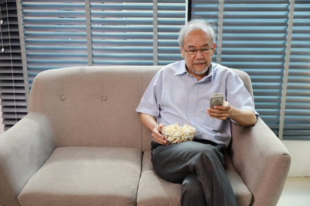 Happy smiling asian senior man sitting on sofa and eating popcorn while having fun with movie rest indoor at home living room. Elderly husband is happiness while watch TVの写真素材