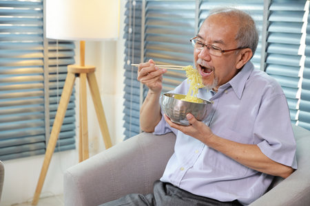 Portrait of asian senior man with beard sitting on sofa eating tasty and instant noodle for lunch in living room with window blinds. Unhealthy and cheap food lifestyle conceptの写真素材