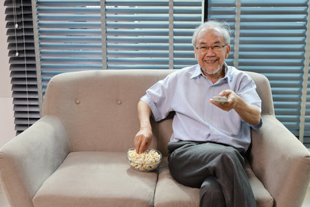 Happy smiling asian senior man sitting on sofa and eating popcorn while having fun with movie rest indoor at home living room. Elderly husband is happiness while watch TVの写真素材