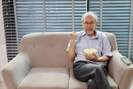 Happy smiling asian senior man sitting on sofa and eating popcorn while having fun with movie rest indoor at home living room. Elderly husband is happiness while watch TVの写真素材
