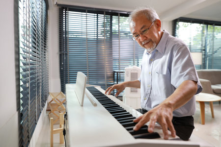 Happy smiling asian senior man with beard sitting and playing piano and singing a song in living room house indorrs. Musical and relaxation makes elder male happiness. Health care lifestyle concept.の写真素材