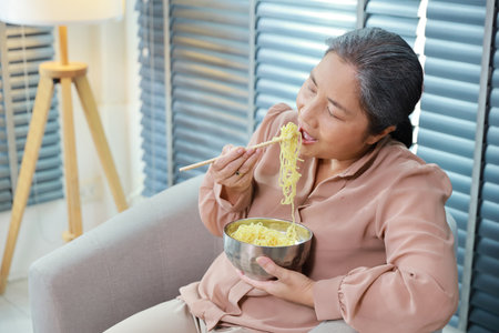 Portrait of asian senior woman with white hair sitting on sofa eating tasty and instant noodle for lunch in living room with window blinds. Unhealthy and cheap food lifestyle conceptの写真素材