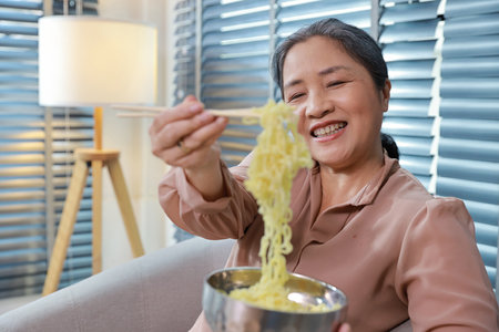 Portrait of asian senior woman with white hair sitting on sofa eating tasty and instant noodle for lunch in living room with window blinds. Unhealthy and cheap food lifestyle conceptの写真素材