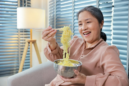 Portrait of asian senior woman with white hair sitting on sofa eating tasty and instant noodle for lunch in living room with window blinds. Unhealthy and cheap food lifestyle conceptの写真素材