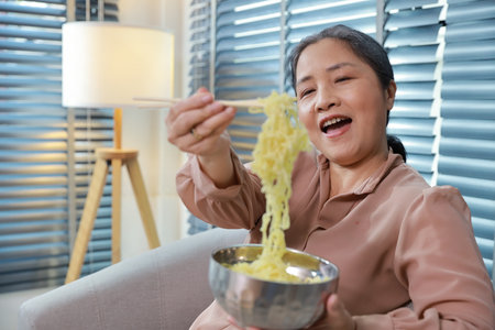 Portrait of asian senior woman with white hair sitting on sofa eating tasty and instant noodle for lunch in living room with window blinds. Unhealthy and cheap food lifestyle conceptの写真素材