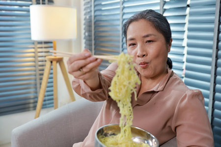 Portrait of asian senior woman with white hair sitting on sofa eating tasty and instant noodle for lunch in living room with window blinds. Unhealthy and cheap food lifestyle conceptの写真素材