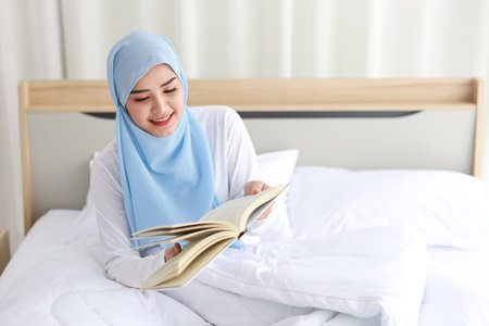 Happy young beautiful asian woman in muslim sleepwear sitting, reading book on bed. Cheerful and cute girl resting in bedroom and copy space for your advertising content.の写真素材