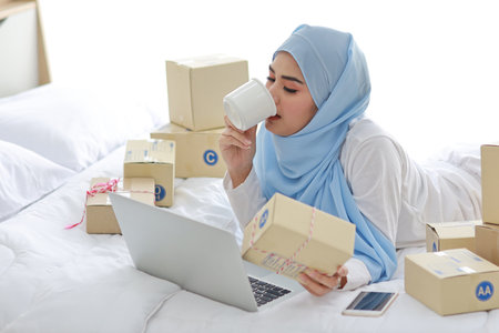 Beautiful and young asian woman in muslim sleepwear with attractive look, lies on bed with computer and online package box delivery. Startup small business SME freelance girl working with mobile phoneの写真素材