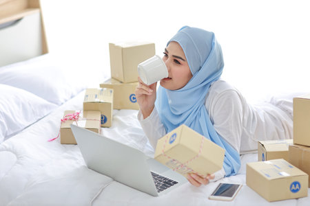 Young attractive asian woman in sleepwear muslim dress, holding coffee cup on bed with computer and online package box delivery. Startup small business SME freelance girl working with mobile phoneの写真素材