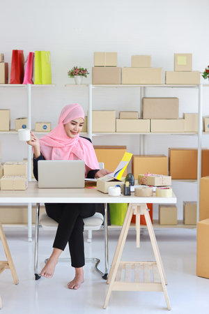 Active smiling asian woman in blue muslim suit sitting and working with online package box delivery. Startup small business SME freelance girl working on computer and mobile phone with happy face.の写真素材