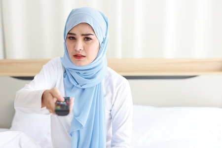 TV and happiness concept. Beautiful asian woman in white muslim sleepwear sitting on bed, holding television remote and watching something with glad happy smiling face.の写真素材