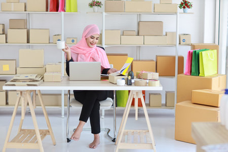 Active smiling asian woman in blue muslim suit sitting and working with online package box delivery. Startup small business SME freelance girl working on computer and mobile phone with happy face.の写真素材