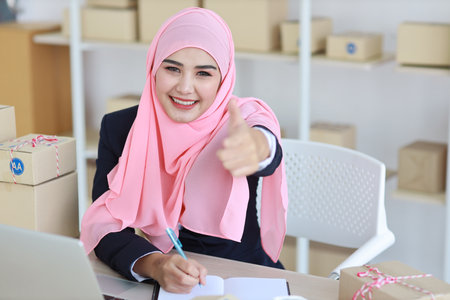 Active asian woman in muslim suit sitting and working with computer and online package box delivery while showing thumb up. Startup small business freelance working girl look at camera with smilingの写真素材