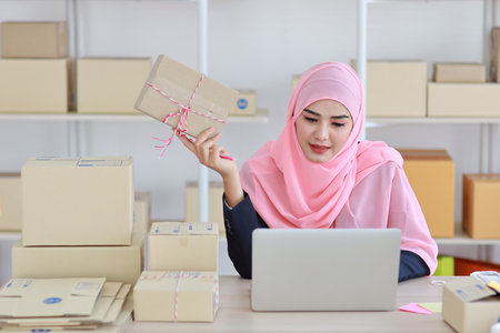 Active asian woman in blue muslim suit sitting and working with computer and showing online package box delivery with happy smiling face. Startup small business freelance working girl look at product.の写真素材