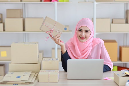 Active asian muslim woman in blue suit sitting and working with computer and showing online package box delivery with happy smiling face. Startup small business freelance working girl look at cameraの写真素材