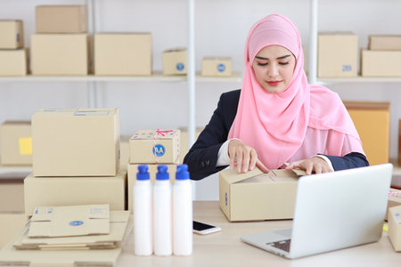 Active smiling asian woman in blue muslim suit sitting and working with online package box delivery. Startup small business SME freelance girl working on computer and mobile phone with happy face.の写真素材