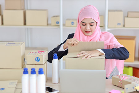 Active smiling asian woman in blue muslim suit sitting and working with online package box delivery. Startup small business SME freelance girl working on computer and mobile phone with happy face.の写真素材