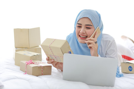 Beautiful and young asian woman in muslim sleepwear with attractive look, lies on bed with computer and online package box delivery. Startup small business SME freelance girl working with mobile phoneの写真素材
