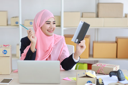 Active asian woman in muslim suit sitting and working with computer, mobile smartphone and online package box delivery. Startup small business freelance working girl checking customer order from bookの写真素材