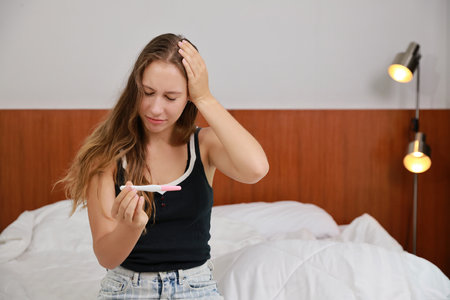 Shocked young woman looking at positive results of pregnancy test. Beautiful girl feeling sad and unexpected for bad news to be future parent on bed in bedroom. Single mon and expecting baby concept.の写真素材