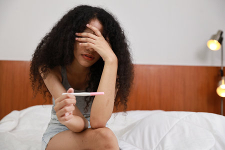 Shocked young woman looking at positive results of pregnancy test. Beautiful girl feeling sad and unexpected for bad news to be future parent on bed in bedroom. Single mon and expecting baby concept.の写真素材