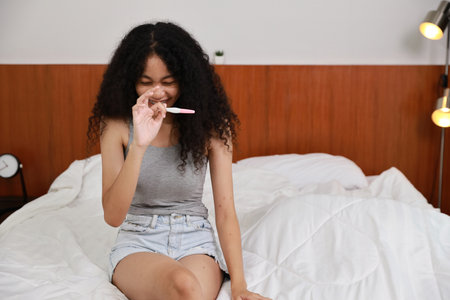 Happy latin young woman looking at positive results of pregnancy test. Beautiful girl feeling happy and excited for good news to be future parent in bedroom. Single mom and expecting baby concept.の写真素材
