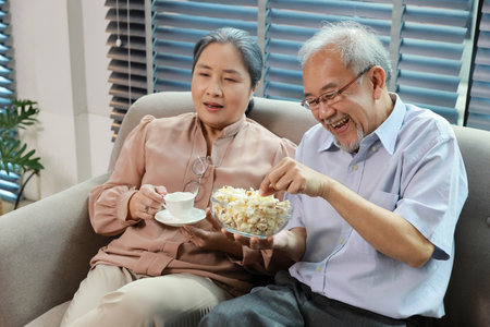 Happy smiling asian senior man and woman sitting on sofa and eating popcorn while having fun with movie rest indoor at home living room. Couple elder husband and wife embrace are happy while watch TVの写真素材